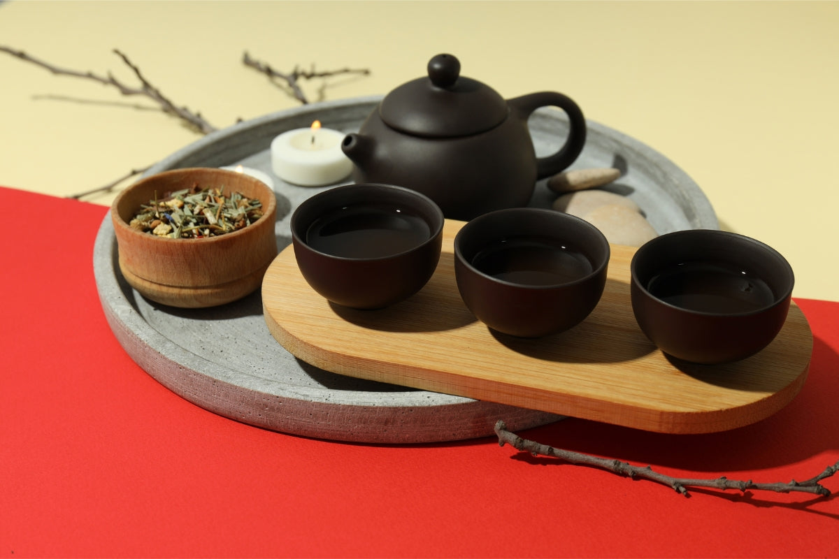 An elegant tea set with a black teapot and three matching cups arranged on a wooden tray, accompanied by a bowl of tea leaves, set against a red and yellow background.