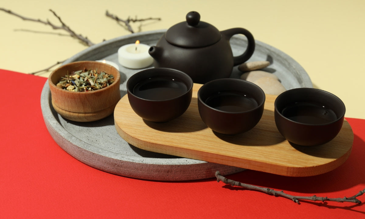 An elegant tea set with a black teapot and three matching cups arranged on a wooden tray, accompanied by a bowl of tea leaves, set against a red and yellow background.